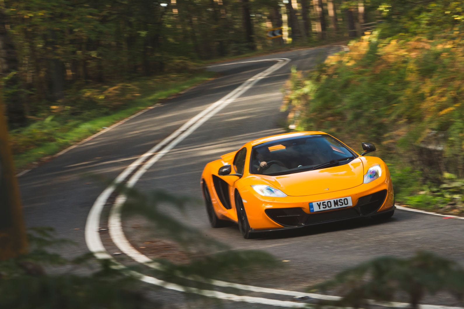 McLaren MP4-12C in orange driving on a winding tree-lined road, illustrating the aerodynamic wing mirror arm design